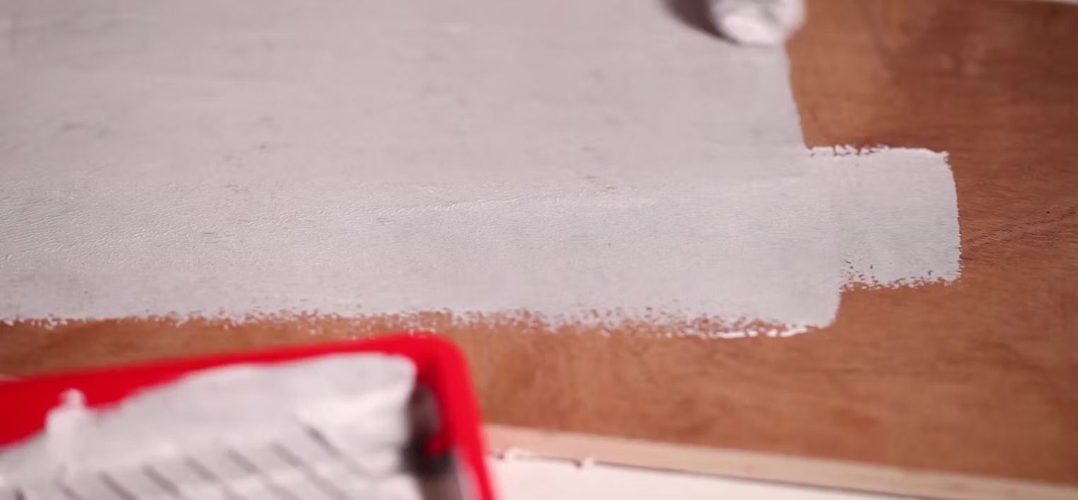 A hand uses a paint roller to apply white primer to a wooden surface next to a red paint tray.