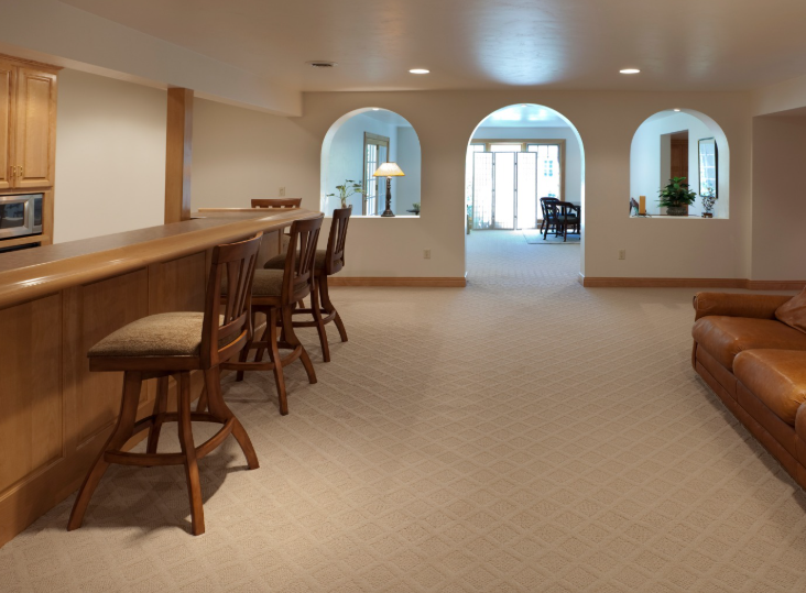 A basement recreation area with a wooden bar, bar stools, arched wall cutouts, and a brown sofa on carpeted flooring.
