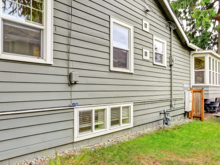 The side exterior of a grey-sided house featuring rectangular windows, a basement window, and a small wooden fence.