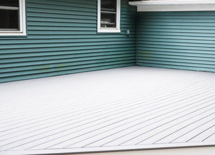 A light gray deck with diagonally laid boards sits against the teal-colored siding of a home with two visible windows.