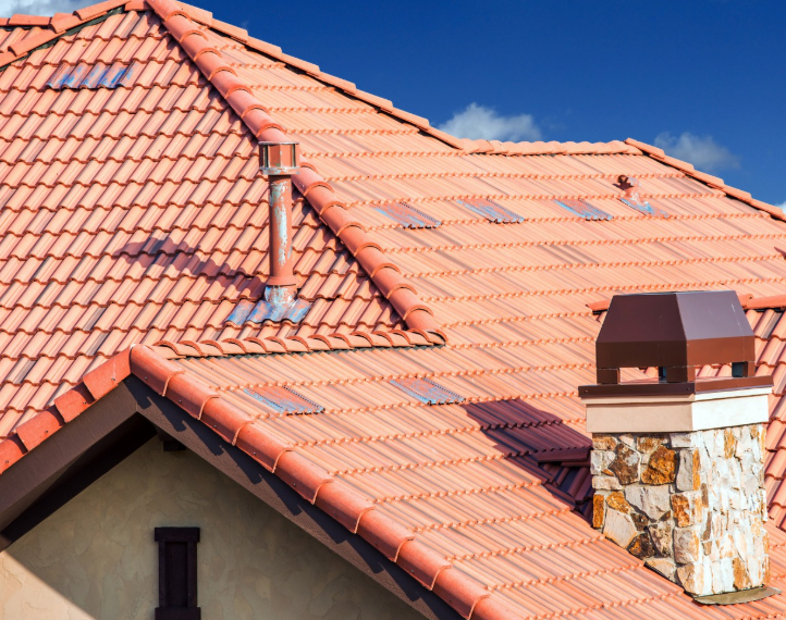 A gabled roof covered in terracotta-colored tiles, featuring a stone chimney, a metal vent pipe, and a blue sky background.