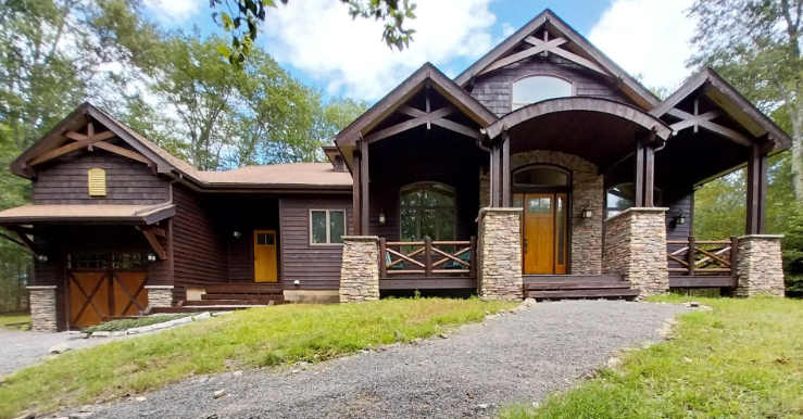 A rustic-style cabin with dark wood siding, a stone foundation, a covered porch, and a gravel driveway in a wooded area.