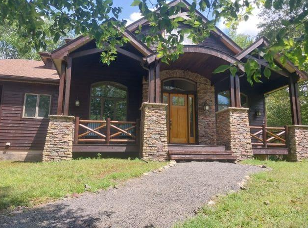 A rustic home with dark brown siding, a covered porch, stone pillars, and a gravel walkway leading to a wooden front door.