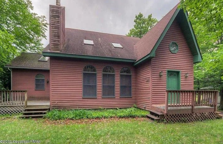 A rustic, reddish-brown house with a steep roof, green trim, and wooden decks, set amidst a lush, tree-filled yard.