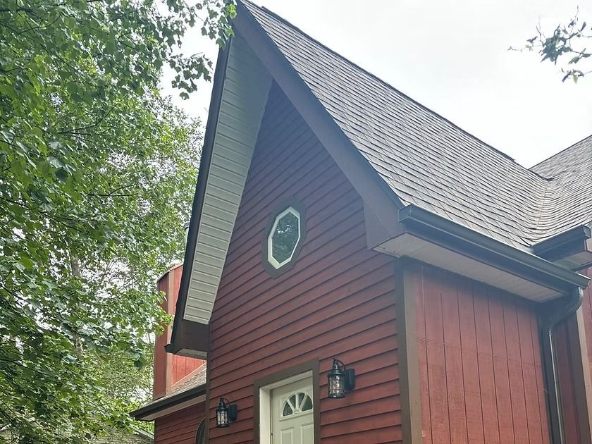 A red house with a steep, pointed gable roof and an oval window above an entrance door, framed by green trees.