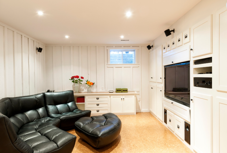 A basement media room with a black sectional sofa, a small window, a white built-in wall unit with a TV, and wood flooring.