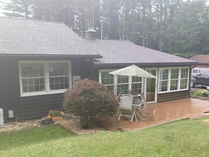 A dark-sided house with a back patio featuring a table, chairs, and an umbrella, set against a wooded backdrop.