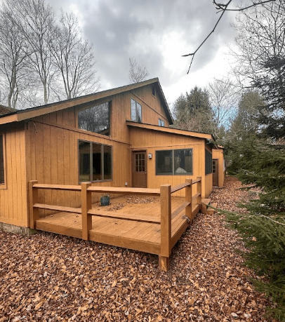 A wooden cabin with a deck, surrounded by a yard covered in fallen autumn leaves under a cloudy sky.