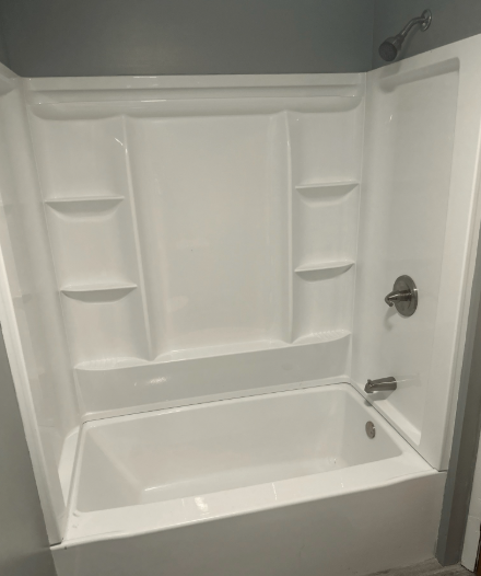 White acrylic bathtub and shower surround with built-in shelves, chrome fixtures, and grey walls.