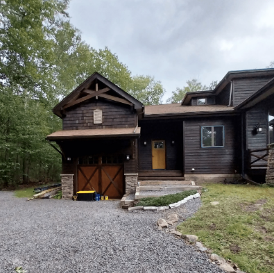 A dark wood-paneled house with a garage, stone accents, and a natural wood door, set in a wooded area with a gravel driveway.