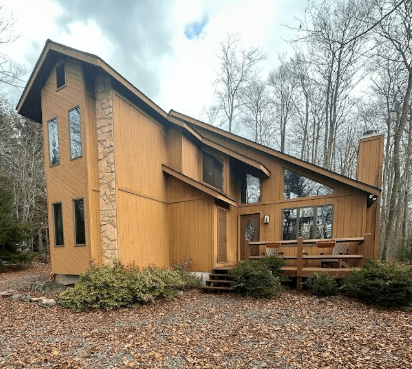 A brown, two-story wooden house with a stone chimney and a small front deck nestled in a wooded, autumn-leaf-covered yard.