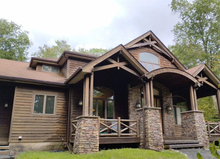 A rustic cabin featuring wood siding, stone pillar supports, a covered front porch, and a peaked roof set among trees.