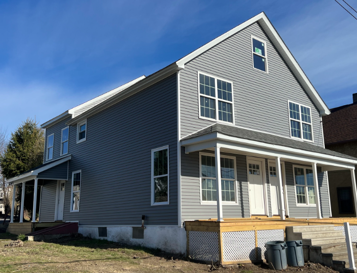 A two-story grey vinyl-sided house with a front porch, white trim, and a small yard under a blue sky.