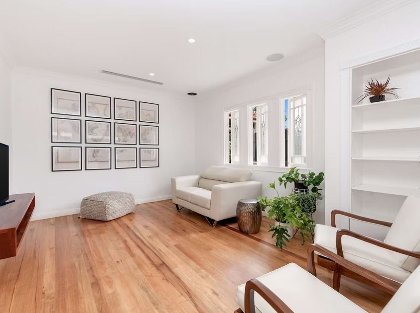 A bright, modern living room with wood floors, white walls, a sofa, a white chair, a shelf, and framed art.