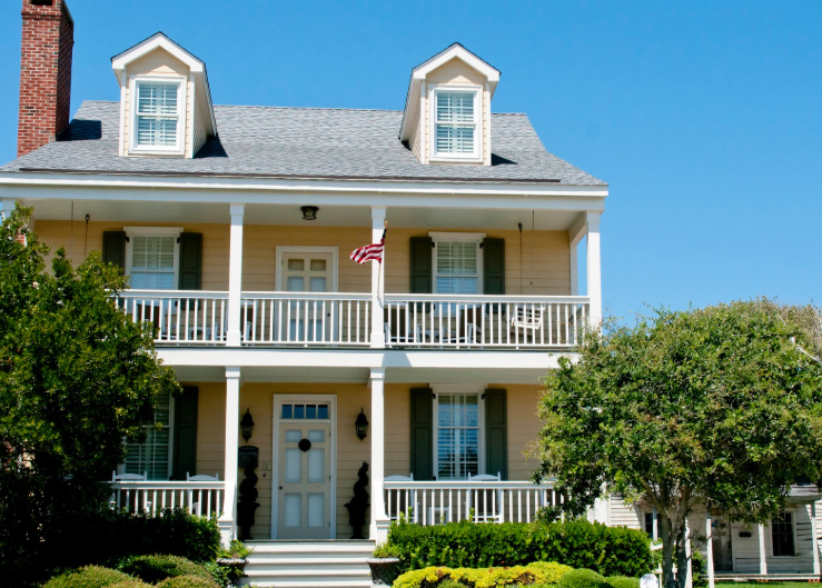 A two-story, yellow colonial-style house with a front porch, white railings, two dormer windows, and a brick chimney.