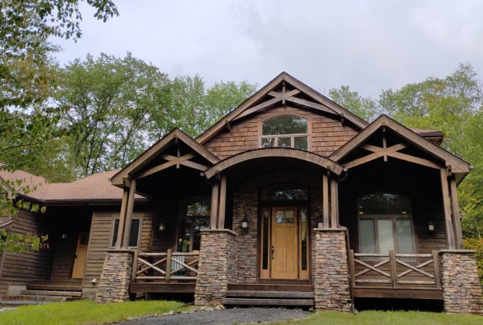 A two-story rustic house with stone pillars, wood siding, and a covered porch, set amidst a forest of trees.