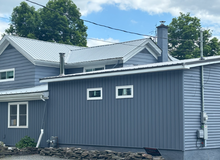A blue-sided house with a white metal roof and a stone foundation against a blue sky with clouds.