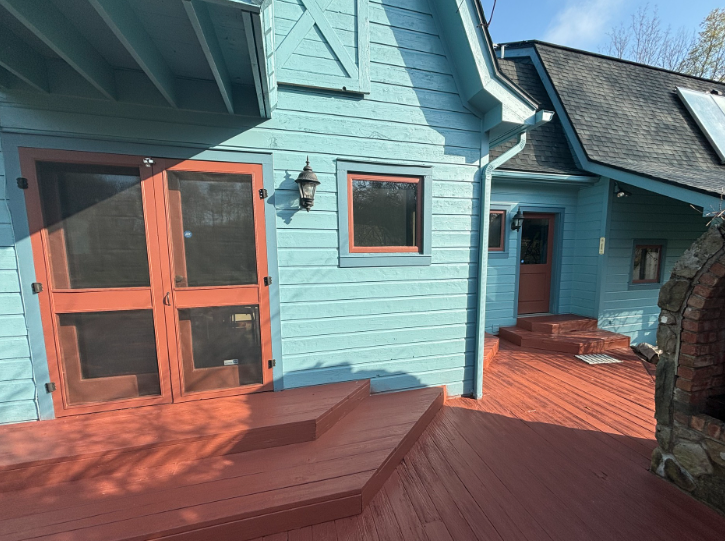 A light blue wooden house with reddish-brown doors and deck stairs, featuring an outdoor stone fireplace area.