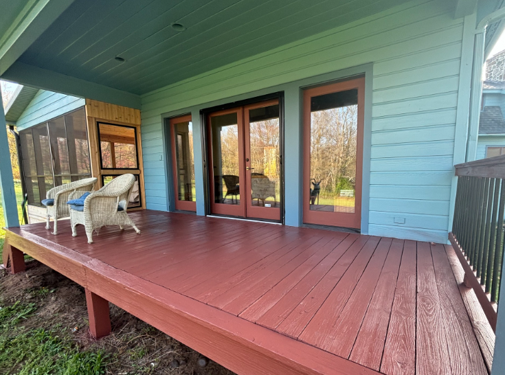 A wooden deck with two wicker chairs sits outside a blue house featuring sliding glass doors and a screened-in porch area.
