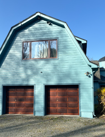 A detached, light blue barn-style garage with a large upper window, shingled roof, and two dark wooden garage doors.