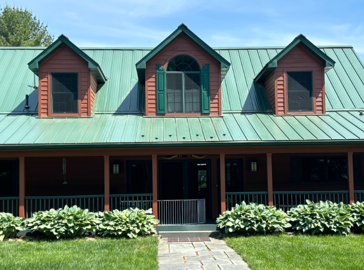 A rustic house with red siding, a green metal roof, three dormers, and a covered porch, surrounded by lush green lawn.