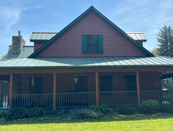 A red-sided cabin with a green metal roof, a full-length front porch with railings, and a stone chimney under a blue sky.