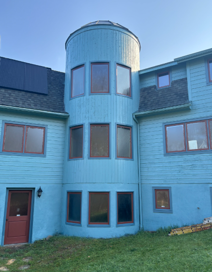 A two-story light blue house with a central, curved turret featuring three vertical rows of windows under a clear sky.
