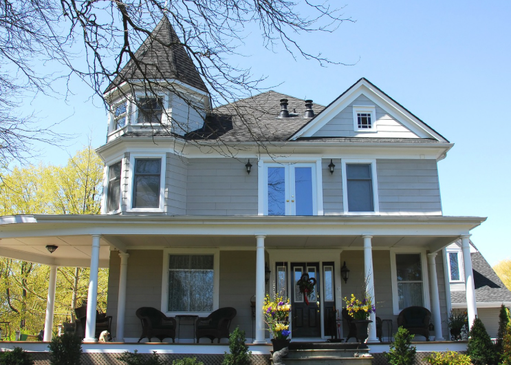 A two-story grey Victorian house with a turret, a wrap-around front porch, and large windows under a clear blue sky.