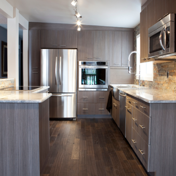 A modern kitchen with wood-tone cabinets, stainless steel appliances, dark wood flooring, and light stone countertops.
