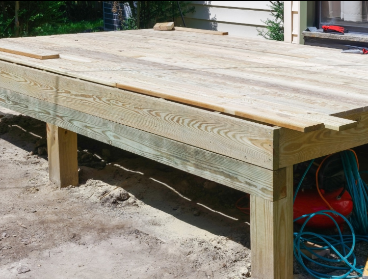 A wooden deck frame under construction on a sandy surface, with tools and a hose stored underneath.