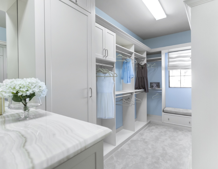 A walk-in closet with white cabinets, light blue walls, a marble-topped vanity with flowers, and a window seat.