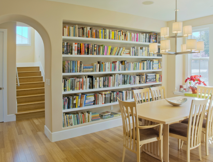 A dining area featuring a built-in wall of bookshelves, light wood flooring, and a modern chandelier above a wood table.