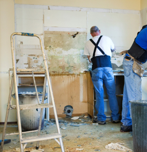 Two construction workers renovate a kitchen wall with a step ladder and debris in the foreground.