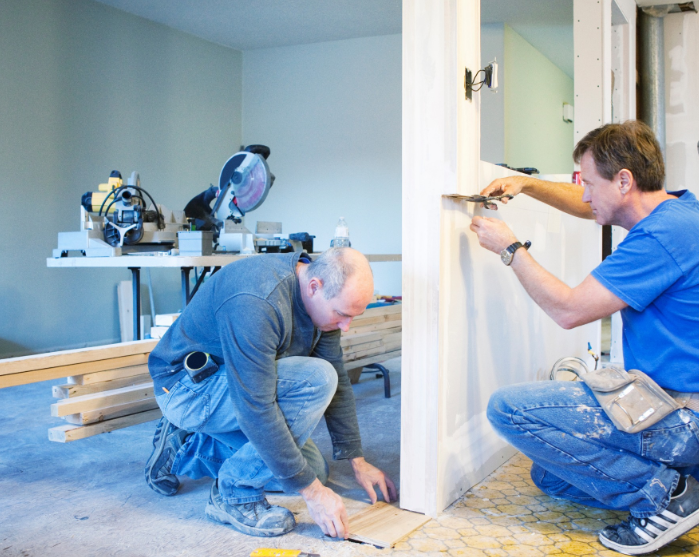 Two contractors working on a home renovation, one installing trim on the floor and the other measuring a wall corner.
