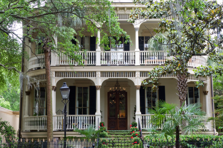 A classic, light-colored two-story Southern house with wrap-around porches, dark shutters, and large trees in front.