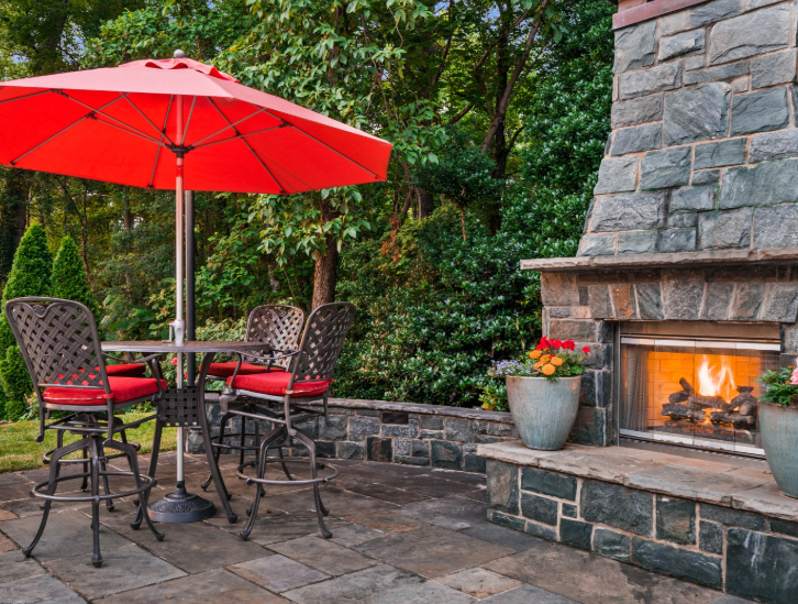 A patio with a stone fireplace, a small table, and chairs under a bright red umbrella in a wooded outdoor setting.