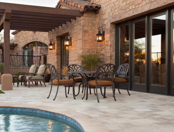 A stone patio beside a swimming pool, featuring a dining set with ornate black metal chairs and a wooden pergola overhead.