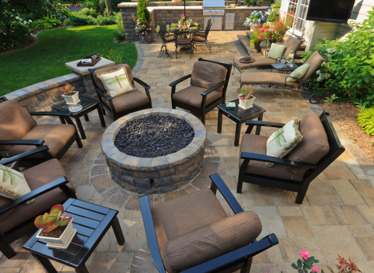 A stone fire pit surrounded by outdoor armchairs and side tables on a paved patio with a dining area in the background.