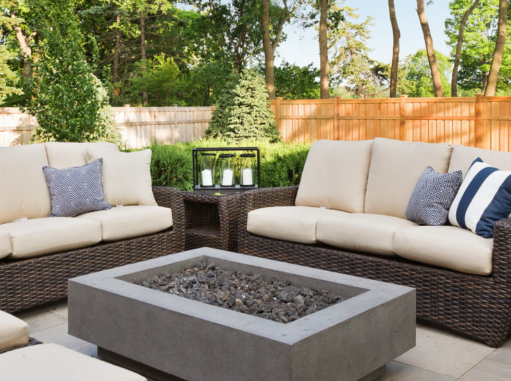Outdoor patio set with two brown wicker sofas and a concrete fire pit on a stone patio with a wood fence background.