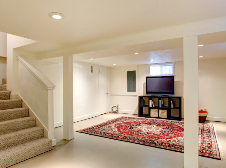 A basement living area with stairs, neutral walls, a flat-screen TV on a media console, and a large red patterned rug.