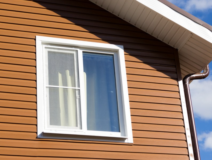 A white window with a screen and a curtain, set in a brown horizontal-sided exterior wall under a roof overhang.