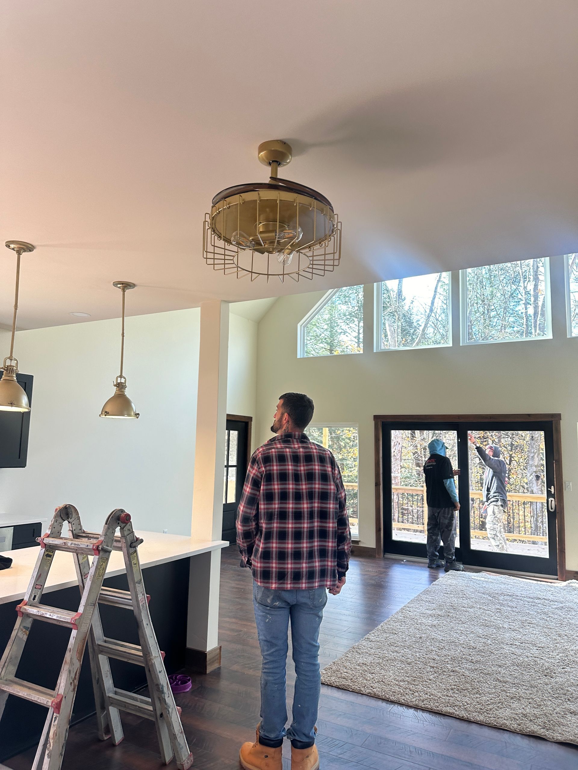 A person in a plaid shirt stands in a room with a decorative ceiling fan, pendant lights, and a sliding glass door.