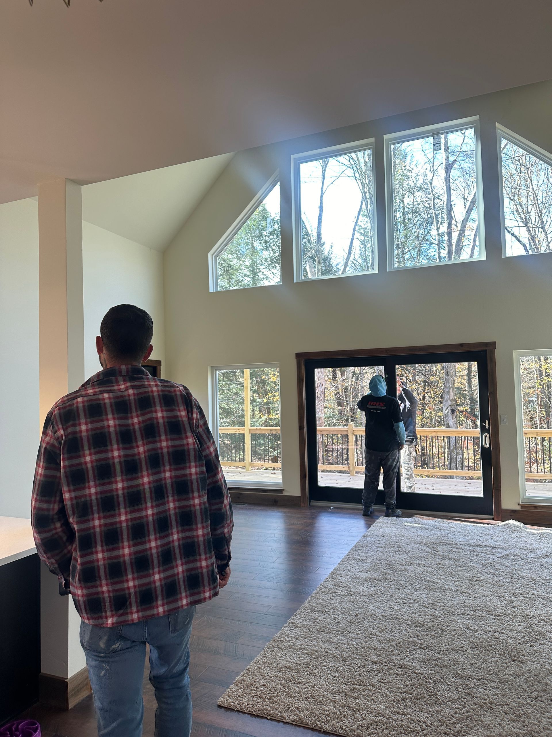 A person in a plaid shirt looks toward a construction worker installing large glass sliding doors in a bright living room.