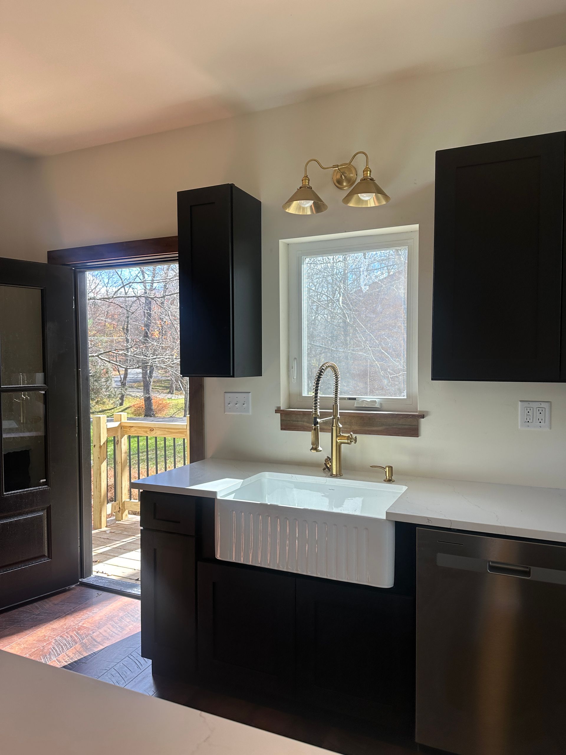 A kitchen sink area with a white farmhouse sink, gold faucet, black cabinets, and an open door to a backyard deck.