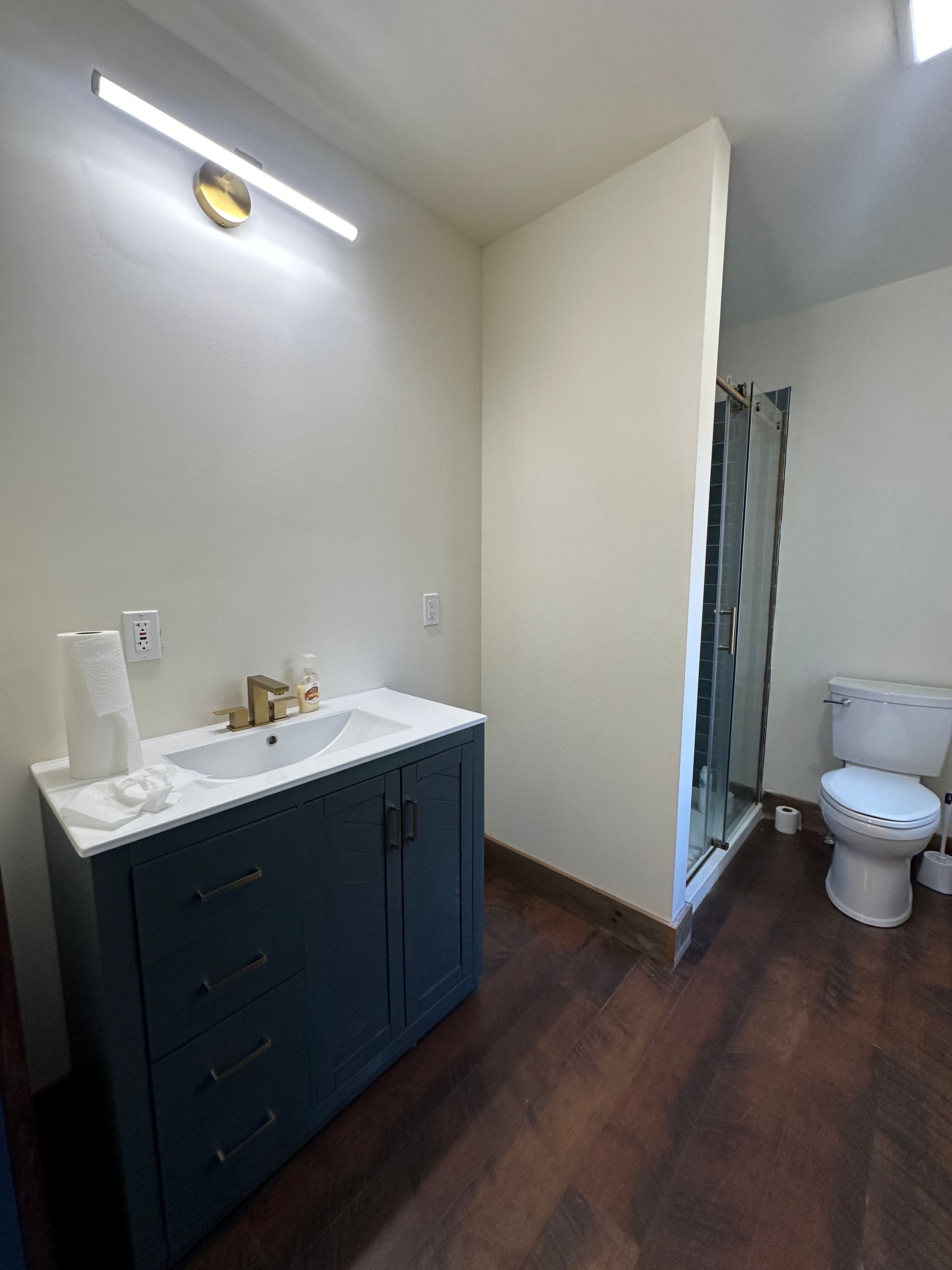 A modern bathroom featuring a dark blue vanity with a white countertop and gold faucet, plus a toilet and shower area.