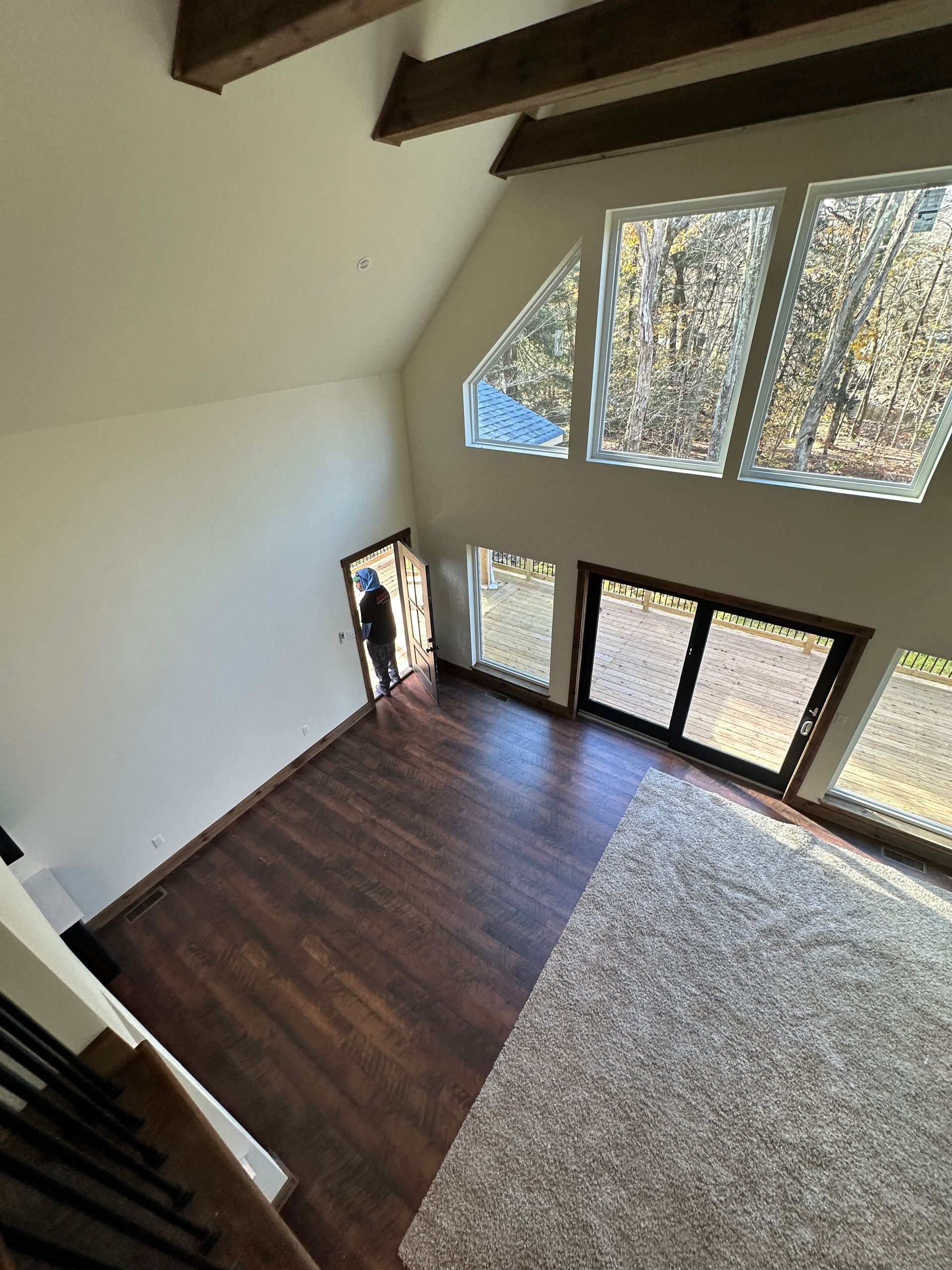 An interior high-angle view of a living room with dark wood flooring, tall windows, exposed beams, and an open doorway.