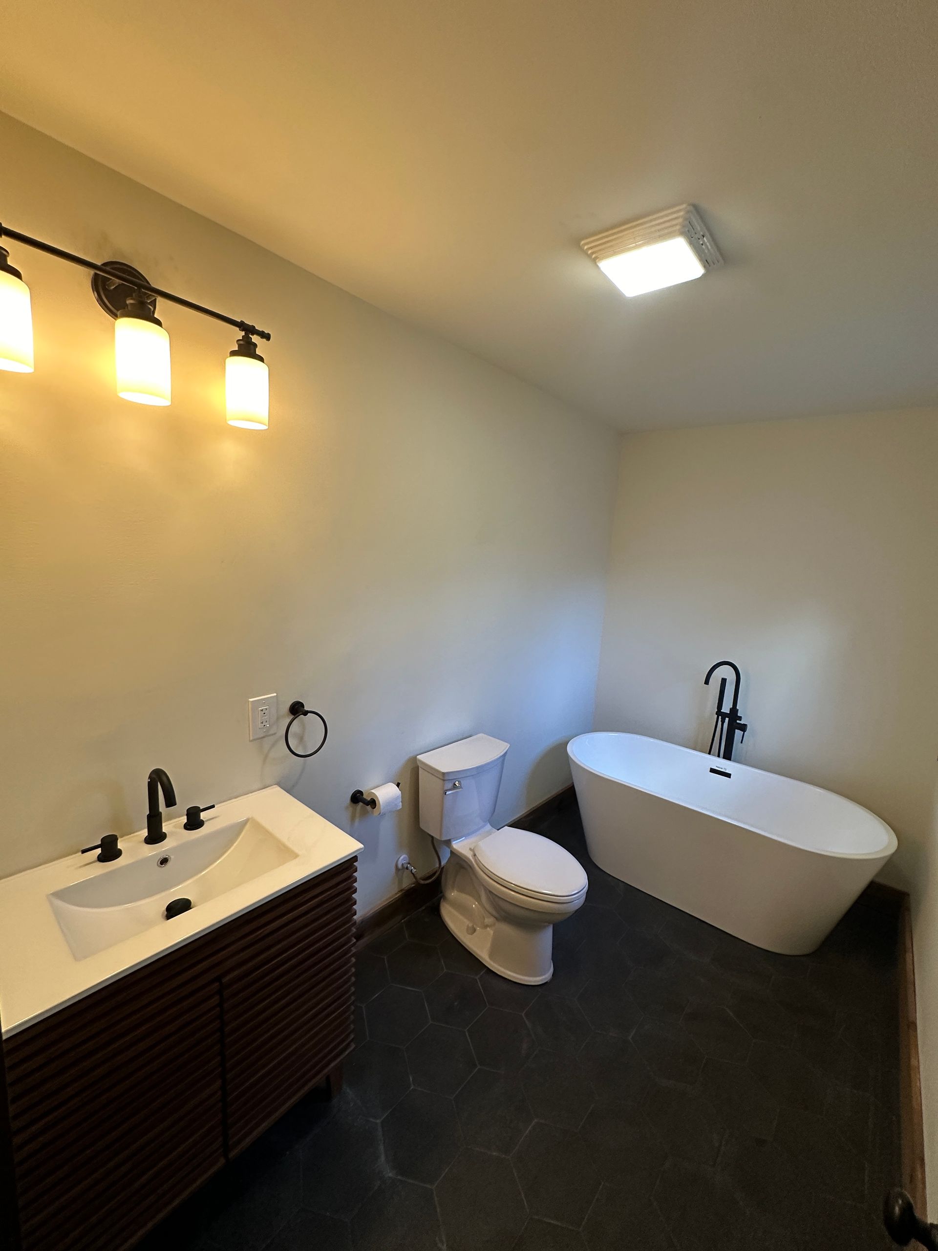 A modern bathroom with a dark wood vanity, a white toilet, and a freestanding white bathtub on a dark tile floor.