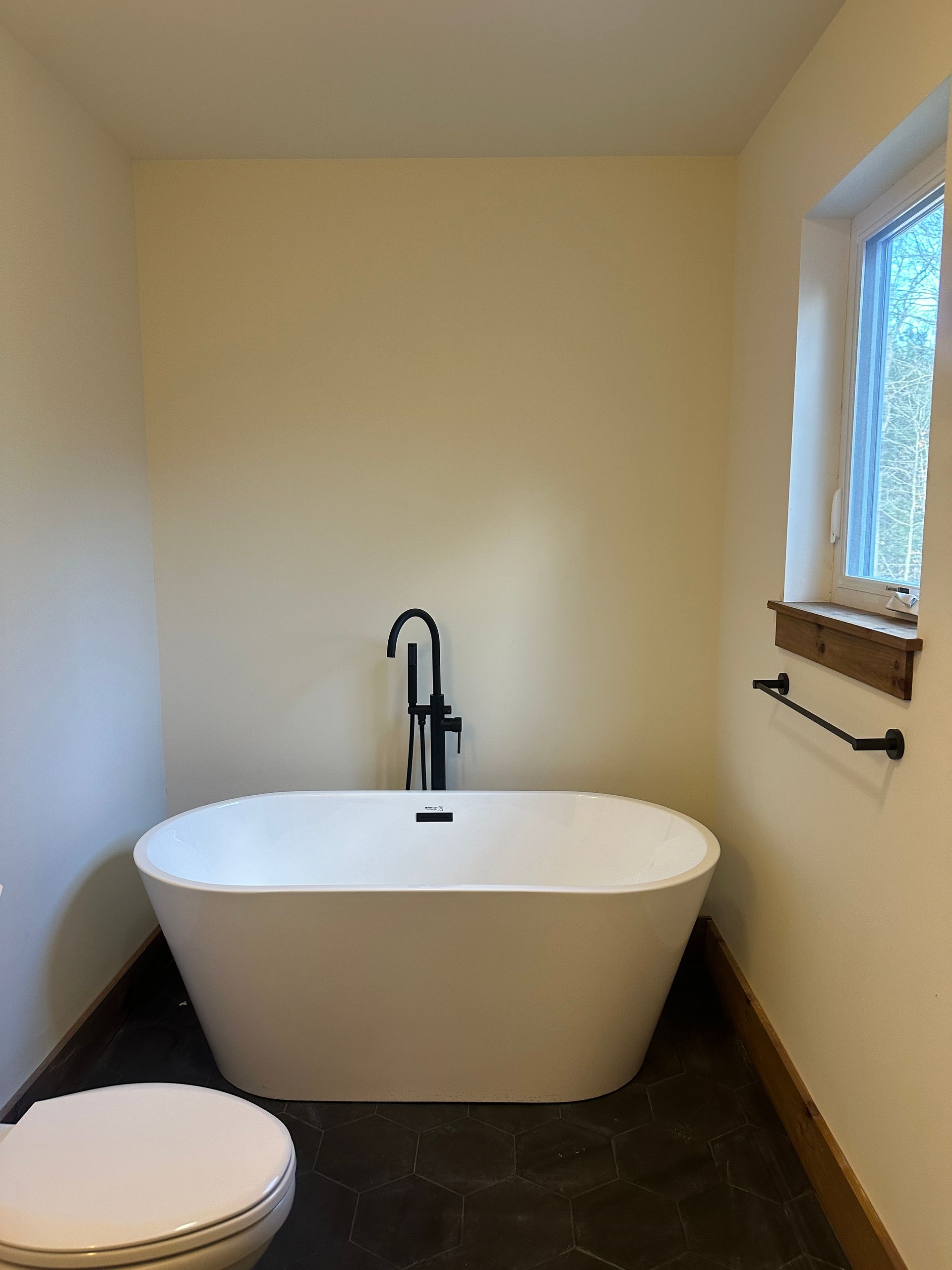 A standalone white bathtub with a black floor-standing faucet in a room with cream walls and a window.