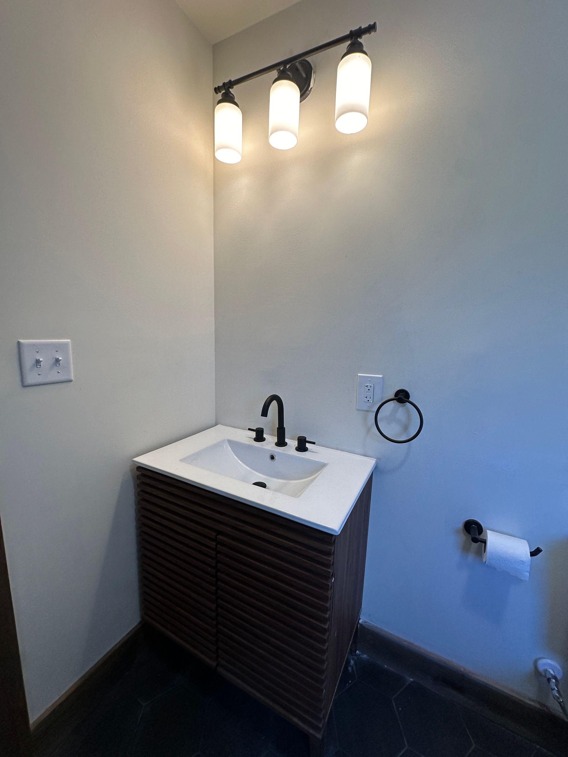 A modern bathroom vanity with a white rectangular sink, dark wood cabinet, and a three-light fixture on a gray wall.