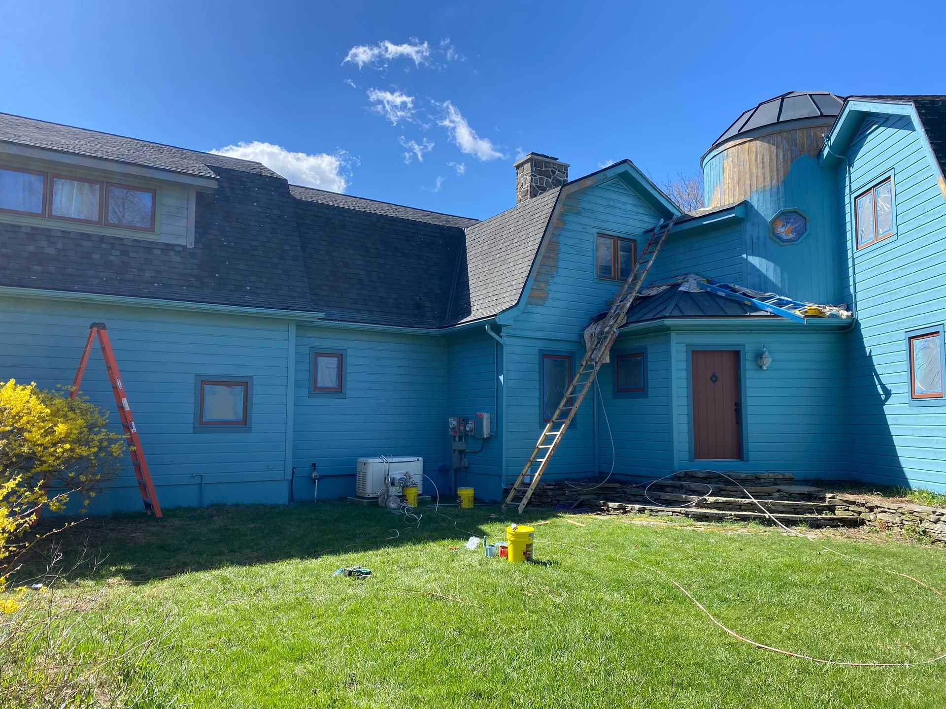 A bright blue, multi-level house with a dome-shaped tower, set against a clear blue sky with a ladder leaning against it.